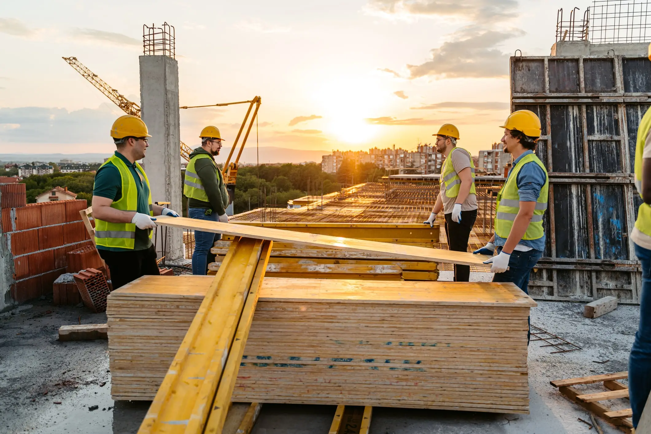 Construction workers in hard hats and hi-vis vests carrying lumber at a job site at sunset